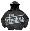 An oversized grey/black hoodie with double zipper, pockets, and a drawstring-free hood, featuring 'The Strokes' text and logo in blue and white across the front body