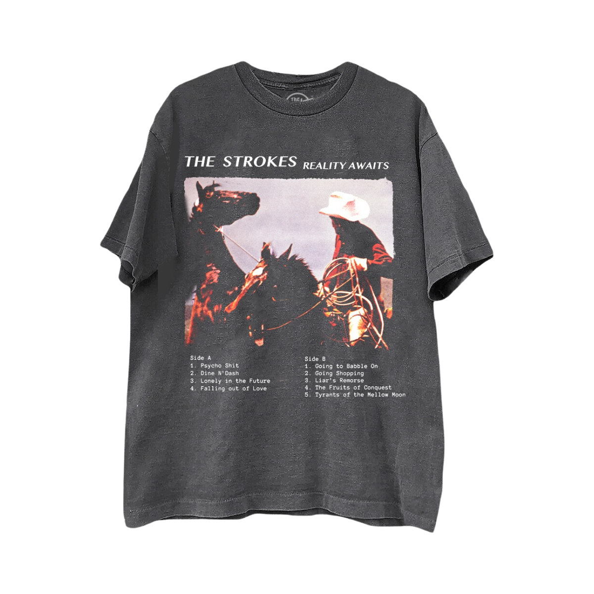 A dark gray graphic t-shirt featuring a western-themed print of a cowboy on horseback with the text 'THE STROKES REALITY AWAITS' on the top and the album tracklist below.