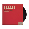The album artwork for The Strokes' Comedown Machine with a black Vinyl poking out. The cover mimics a vintage RCA magnetic tape box, featuring a bold, textured red background and a classic, cream-colored RCA manufacturer label across the top. The album title is stamped in a minimalist, black sans-serif font across the center, giving the packaging a retro, analog media aesthetic.